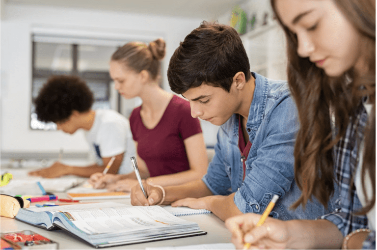 Students sitting at desks in a classroom, focused on writing in notebooks and studying from textbooks.