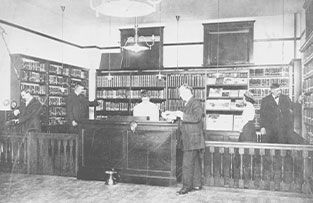A black-and-white historical photo of a library interior with staff behind a large service desk and shelves of books.