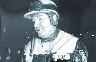 A black-and-white portrait of a race car driver wearing a helmet and goggles, smiling slightly in a dark setting.