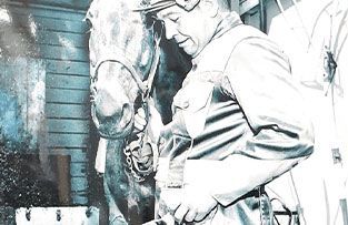 A black-and-white, slightly blurred photo of a person in a uniform standing beside a horse in a stall.