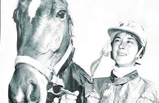 A jockey wearing a helmet and racing silks standing beside a horse with a white marking on its forehead.