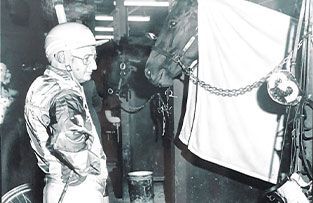 A jockey wearing a helmet and racing silks stands in a stable, looking at a horse wearing a blanket with a number three.