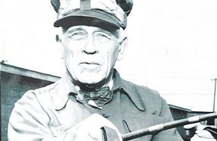 A person in a military-style uniform and cap holding a baton or pointer, looking forward in a black-and-white photograph.