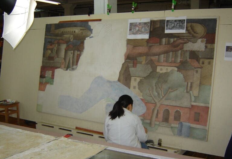 A person in a white lab coat restores a large, partially painted mural featuring a building, trees, and an airplane.