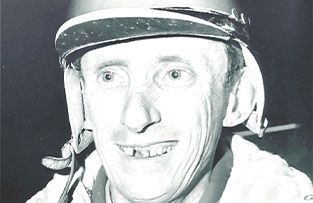 A smiling jockey wearing a protective helmet, captured in a close-up, black-and-white portrait.