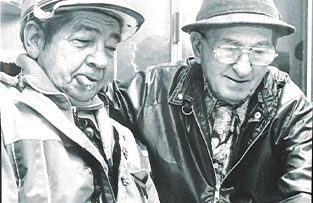 Two people side by side; one wearing a cap and jacket, the other wearing a hat, glasses, and a jacket with a hand on the shoulder.