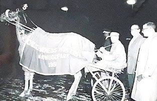 A horse wearing a Maywood Park 1957 blanket pulls a sulky with a driver, flanked by two men standing on a racetrack.