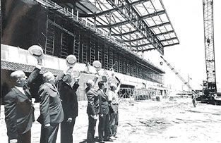 A group of men in suits raising their hats toward the steel framework of a large stadium under construction.