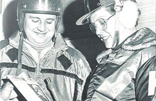 Two people wearing winter gear and headwear look down at a document while standing close together.