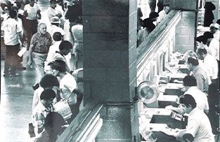 A black-and-white view of workers standing at a long service counter, interacting with staff using equipment.