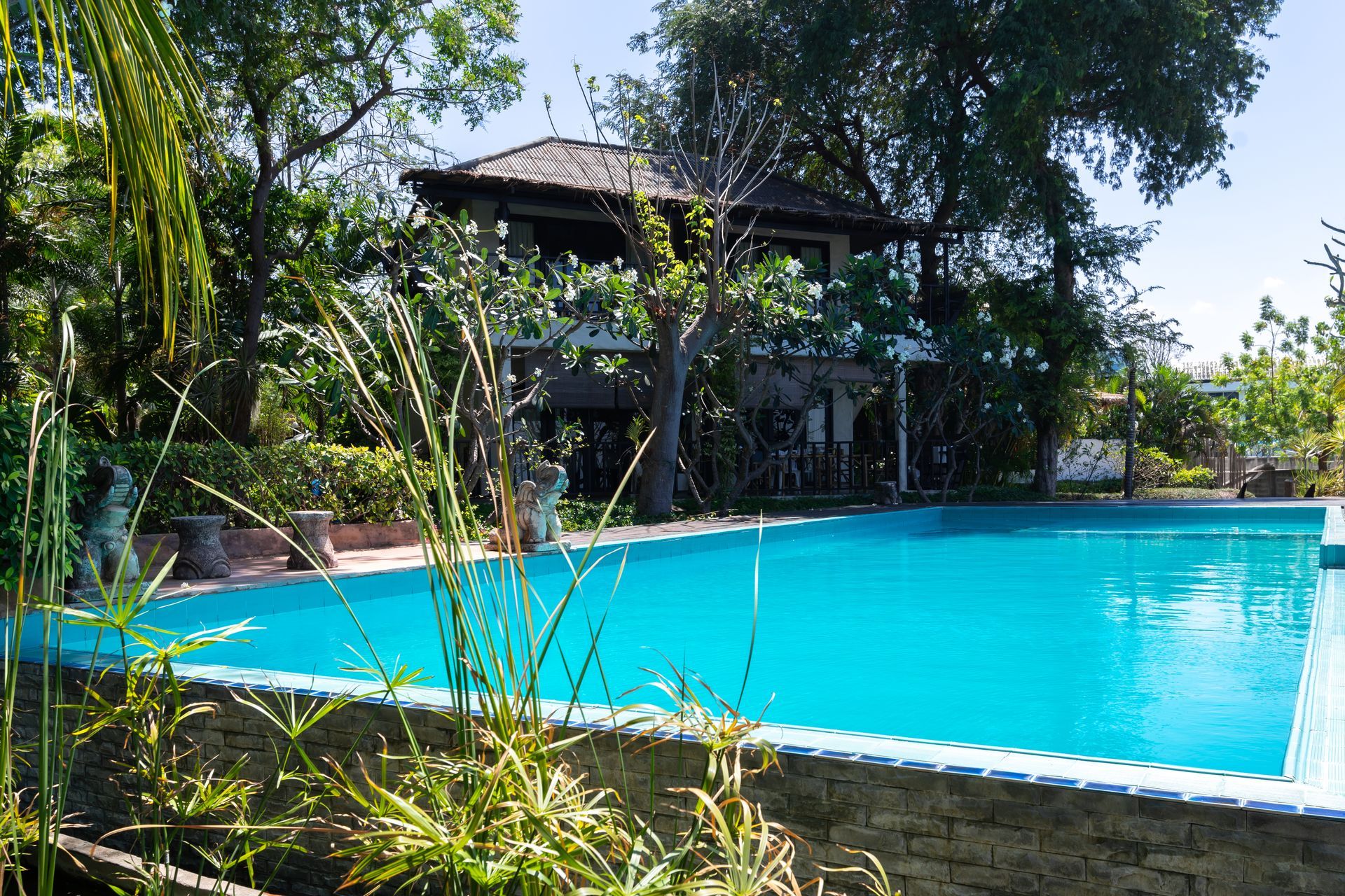 A bright blue swimming pool sits in front of a two-story building surrounded by lush green trees on a sunny day.