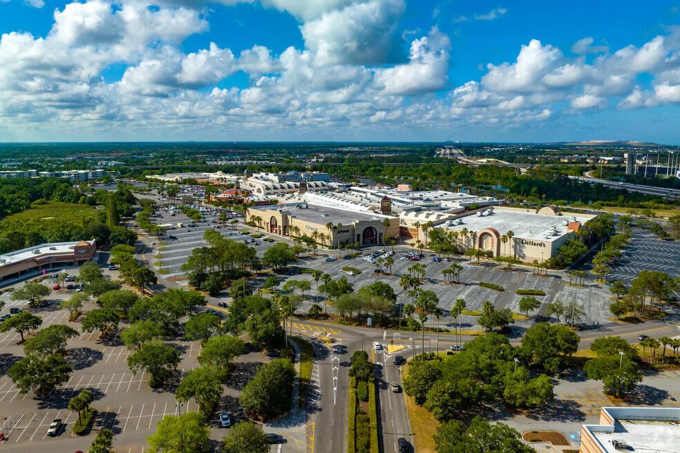 Aerial view of a large shopping mall complex surrounded by extensive parking lots, greenery, and trees under a blue sky.