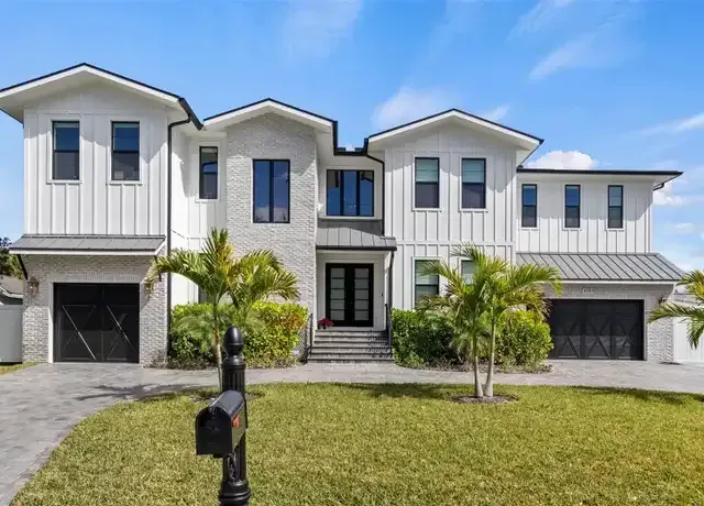 A modern two-story white house with two garages, stone accents, and palm trees in a grassy front yard.