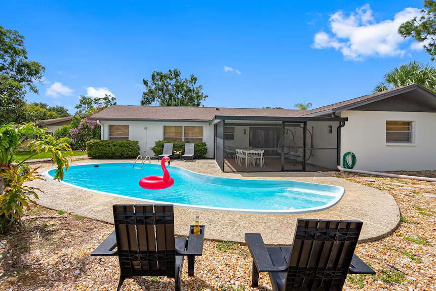 A backyard swimming pool with a pink flamingo float, two lounge chairs in the foreground, and a house with a patio.