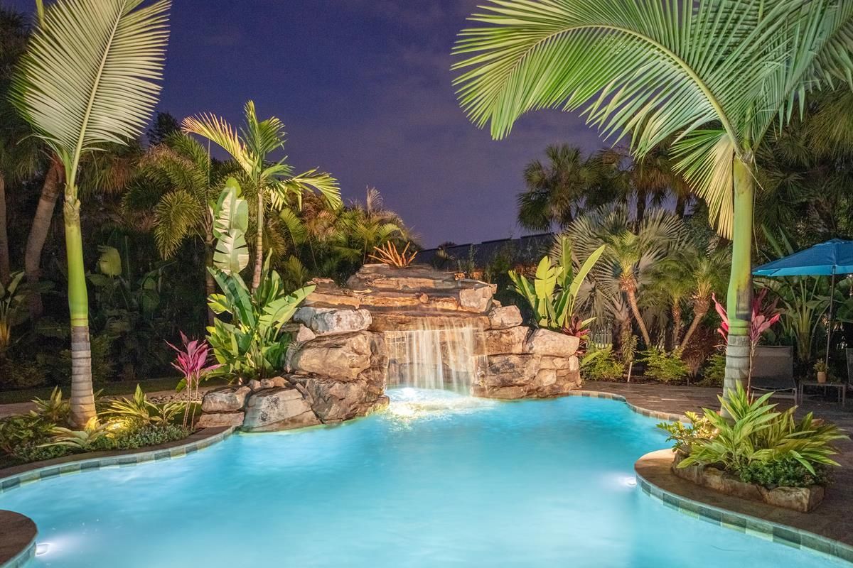 A tropical-style swimming pool at night, featuring a stone waterfall, glowing blue water, and lush palm trees.