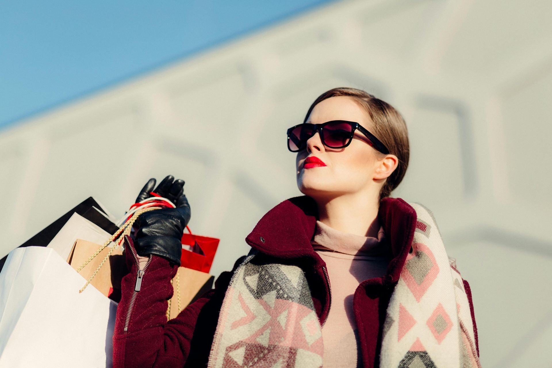 Woman in sunglasses holding shopping bags, looking up. Wearing coat and scarf, bright sunny day.