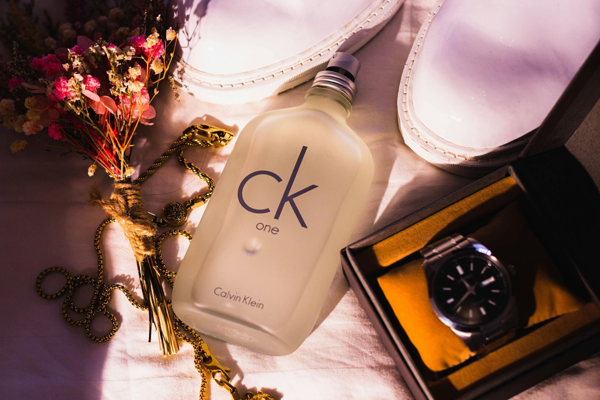 CK One cologne bottle, watch in box, and shoes on a white surface with dried flowers and gold necklace.