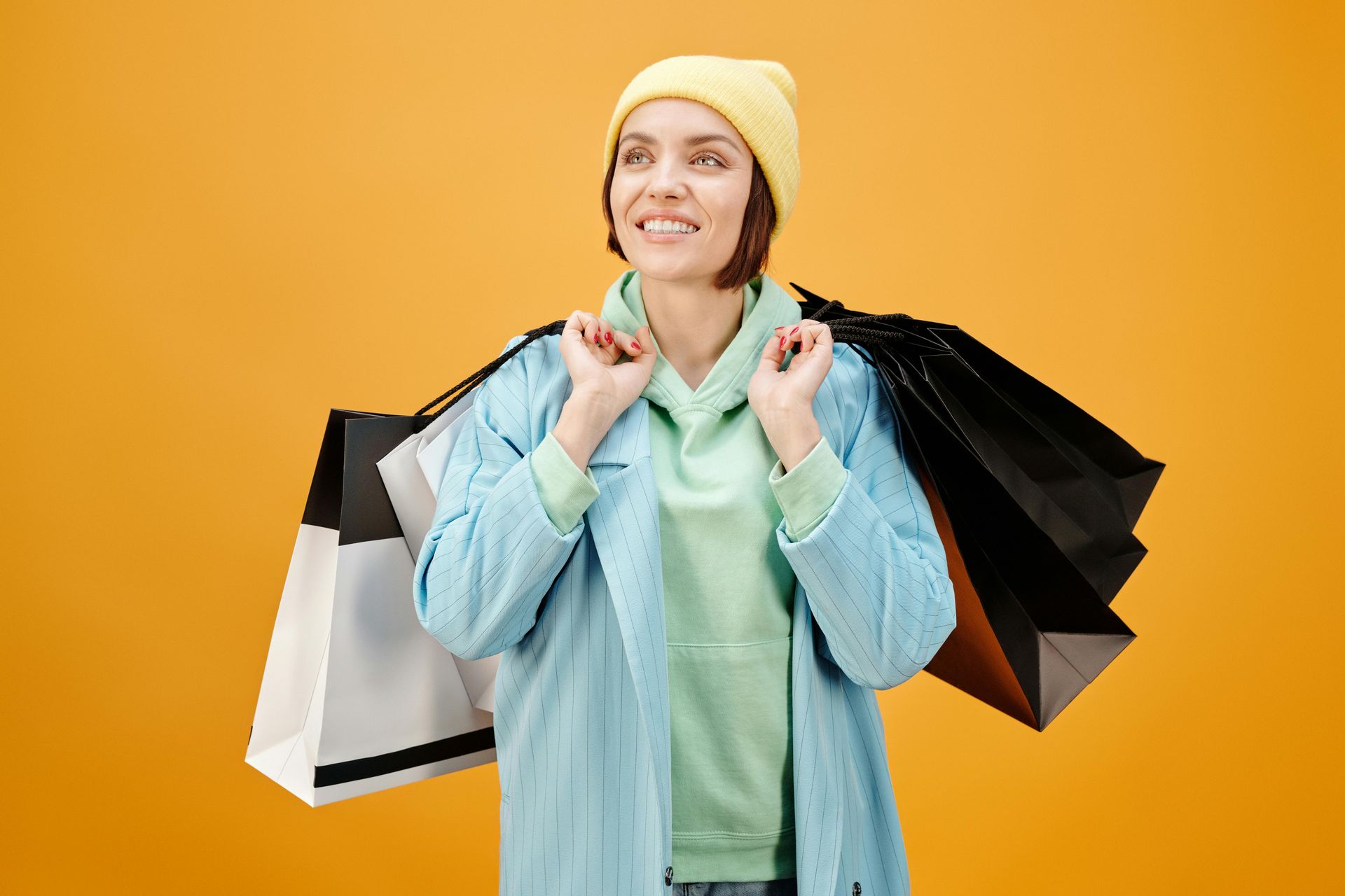 Woman holding shopping bags, smiling, wearing a blue coat and yellow hat against a yellow background.