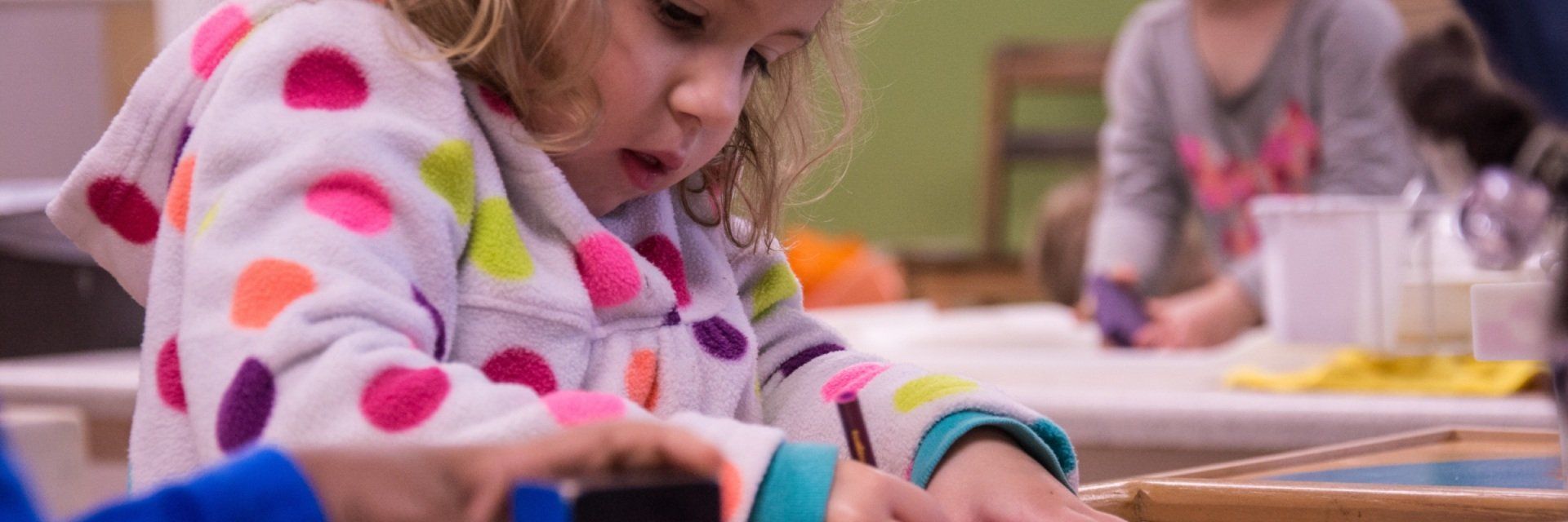 A little girl in a colorful polka dot shirt is playing with a box.