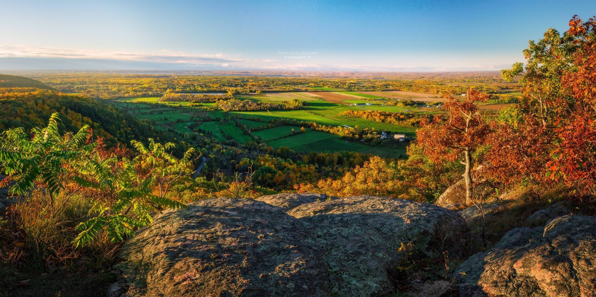 Autumnal landscape from a rocky overlook with colorful foliage and a vast green valley under a blue sky.