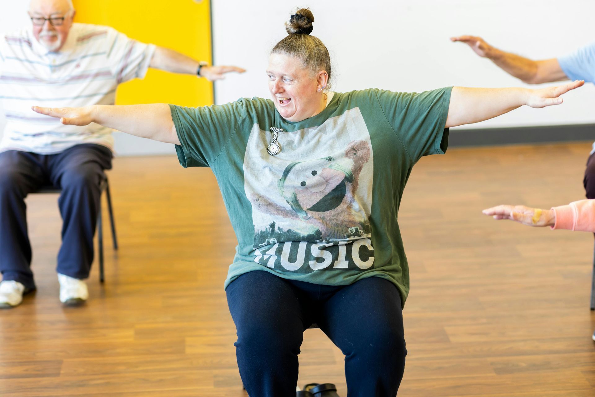 Person seated, arms outstretched, exercising in a room with other seated individuals.