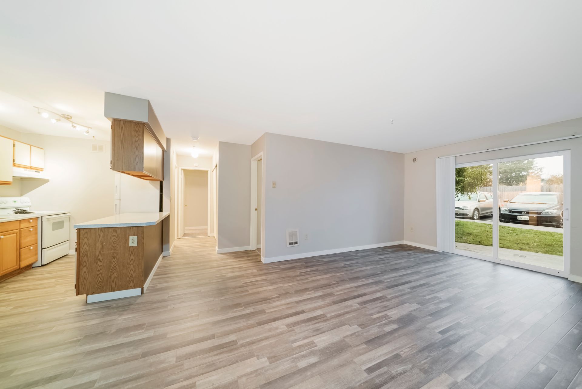 An empty living room with hardwood floors and sliding glass doors leading to a kitchen.