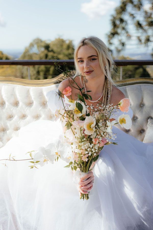 A Woman in a Wedding Dress is Sitting on a Couch Holding a Bouquet of Flowers — Prim n Proper Event Hire in Palmview, QLD