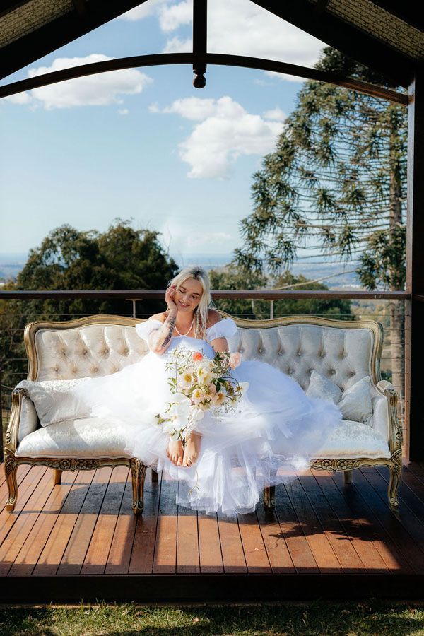 A Bride in a White Dress is Sitting on a Couch Holding a Bouquet of Flowers — Prim n Proper Event Hire in Palmview, QLD