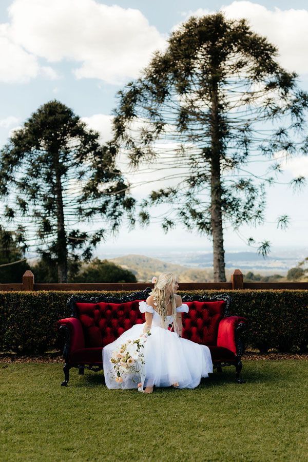 A Bride in a White Dress is Sitting on a Red Couch in the Grass — Prim n Proper Event Hire in Palmview, QLD
