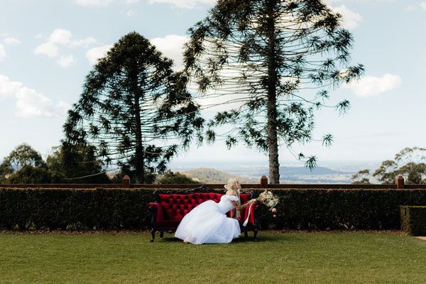 A Bride in a White Dress is Sitting on a Red Couch in the Grass — Prim n Proper Event Hire in Palmview, QLD
