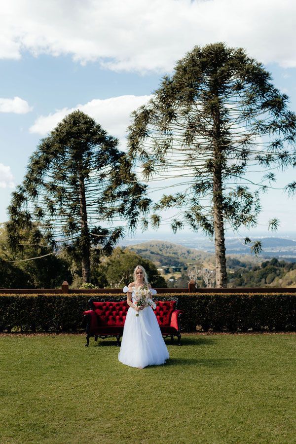 A Bride in a White Dress is Standing in a Grassy Field Next to a Red Bench — Prim n Proper Event Hire in Palmview, QLD