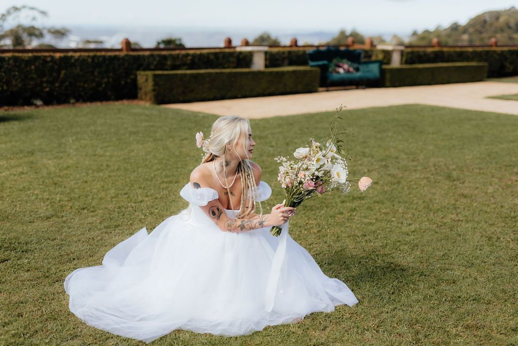 A Bride in a Wedding Dress is Sitting on the Grass Holding a Bouquet of Flowers — Prim n Proper Event Hire in Palmview, QLD