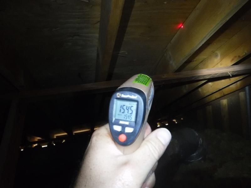 Hand holding infrared thermometer in an attic, displaying 154.5. Laser dot on wooden beam. Dark setting.