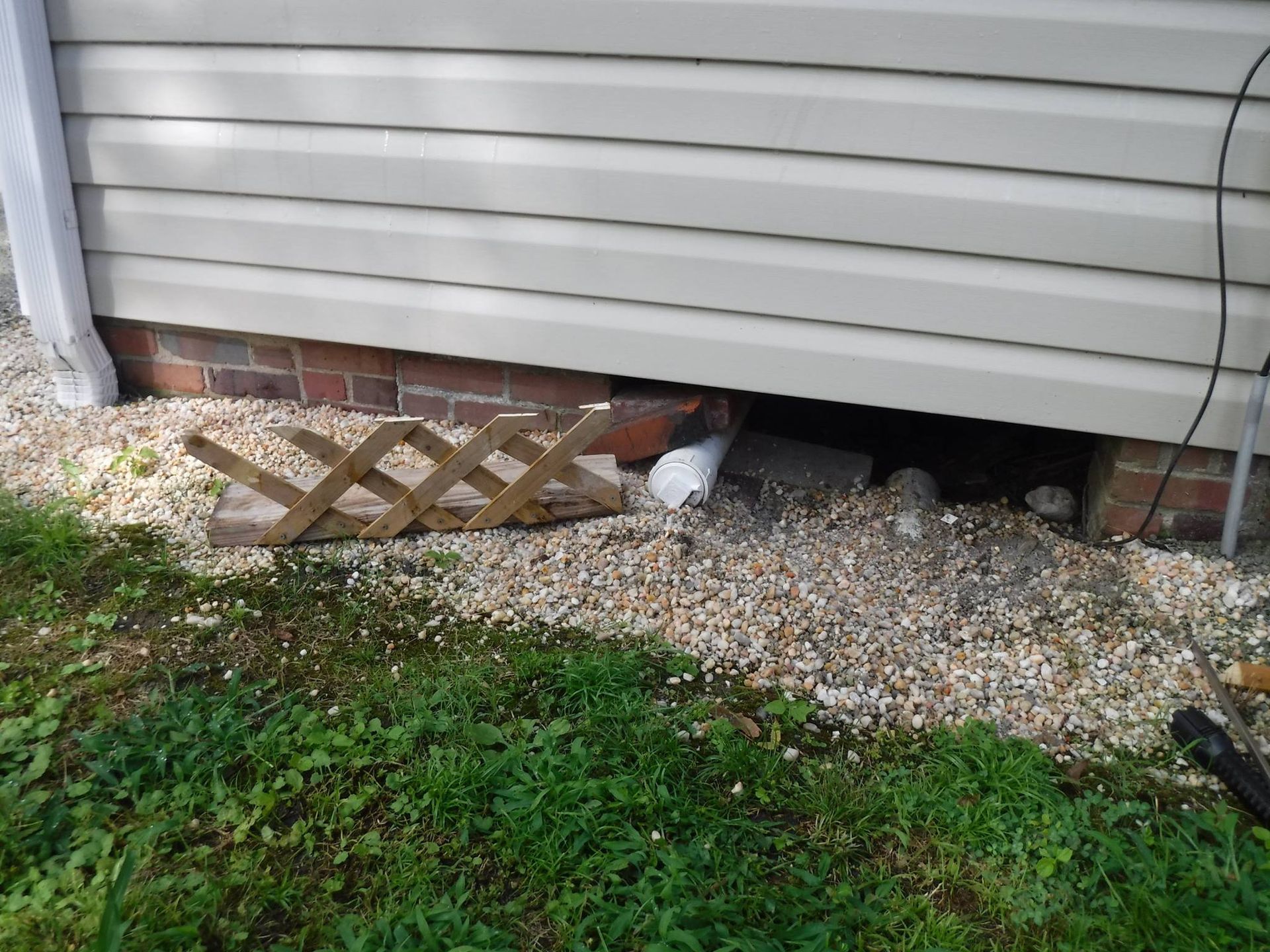 Lattice fence against a house with tan siding, exposed brick foundation, and gravel, next to green grass.