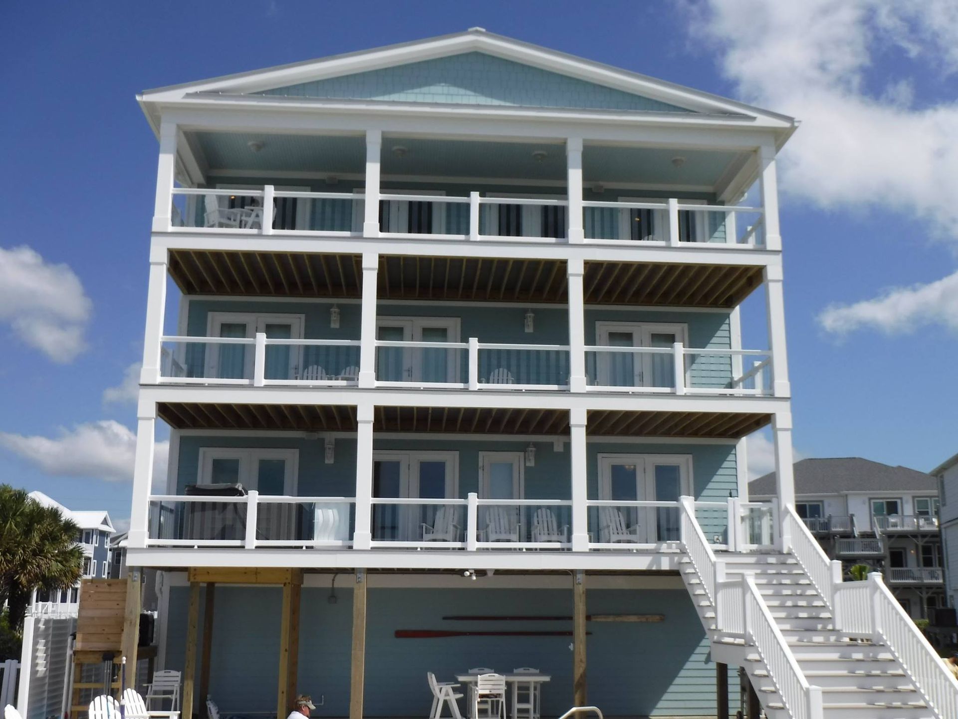 Blue three-story beach house with balconies, white stairs, and a bright blue sky.