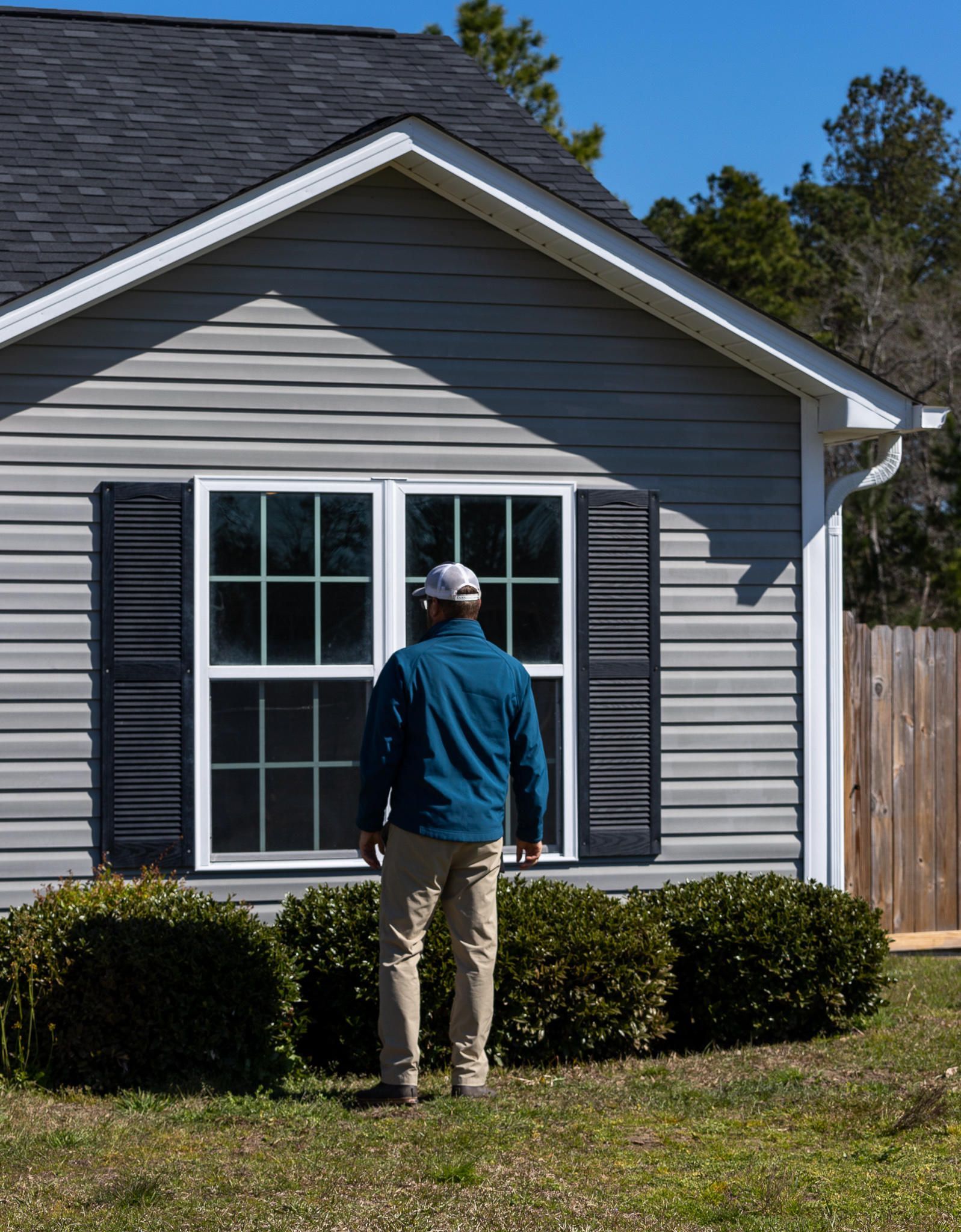Person standing in front of a house, looking at the window. The house has gray siding and black shutters.