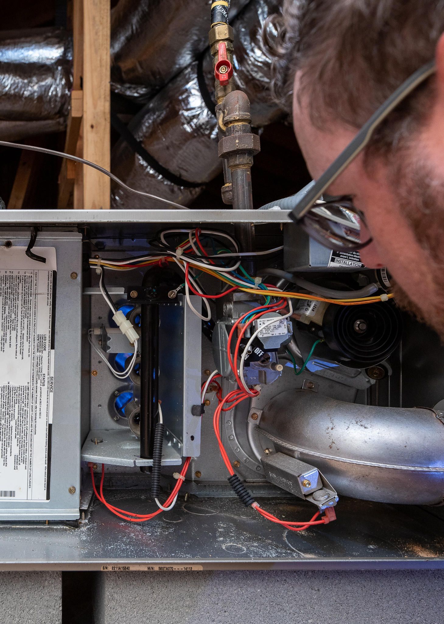 Man inspecting furnace wiring, wearing glasses, in a mechanical room; wires and metal components visible.