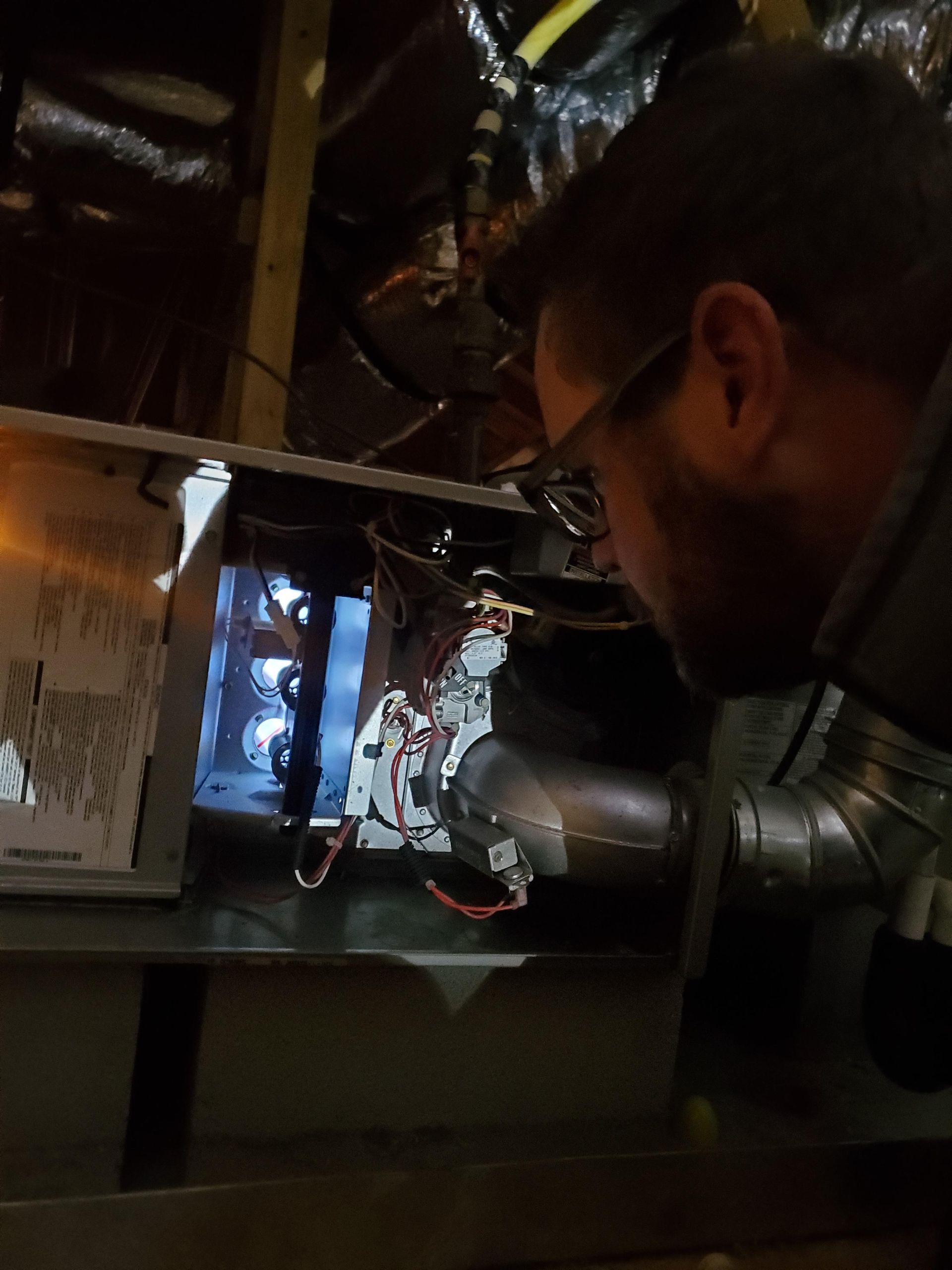 Man examining furnace components with a flashlight in a dark, confined space.