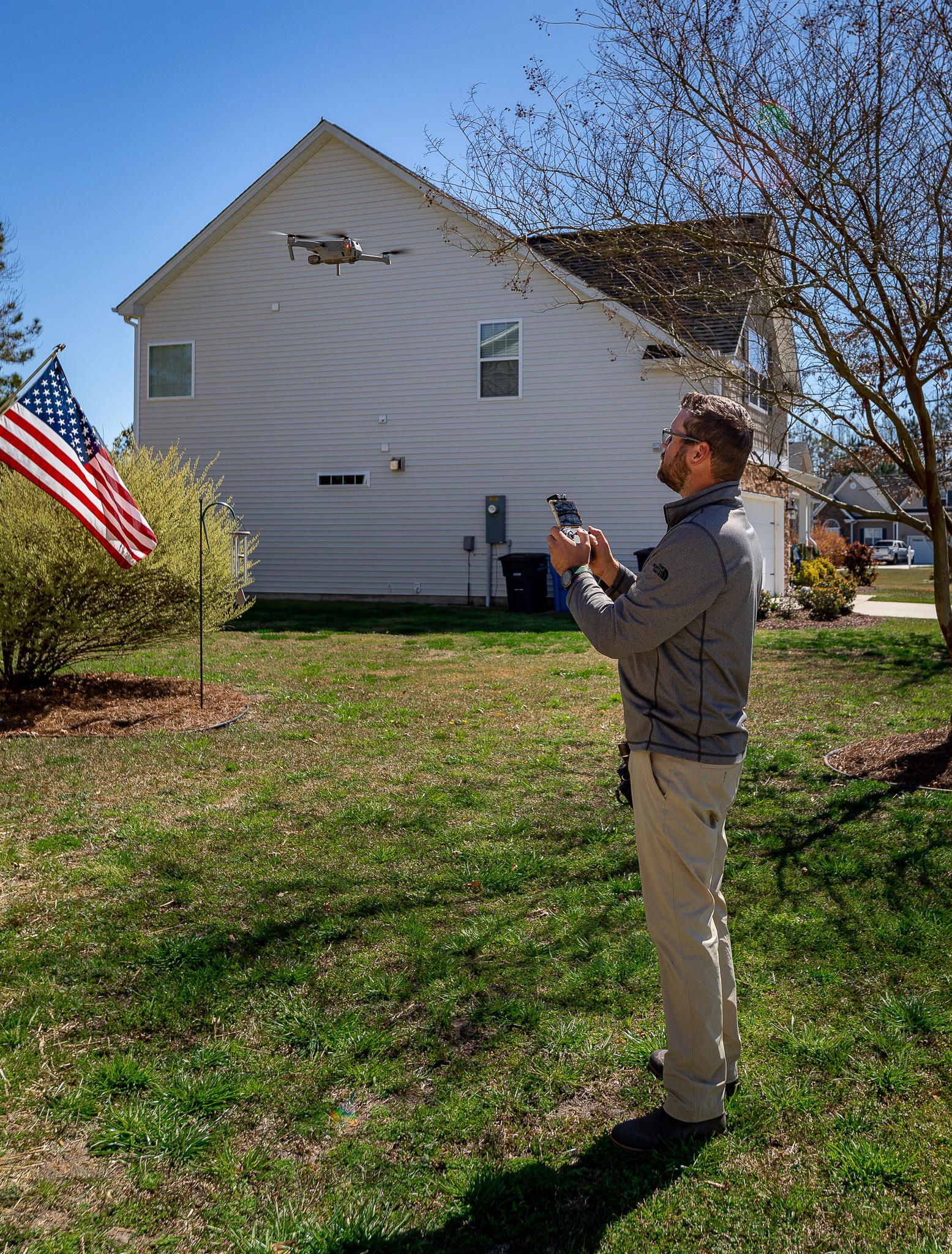 Man operating a drone in a grassy yard. White house, American flag, and tree in the background.