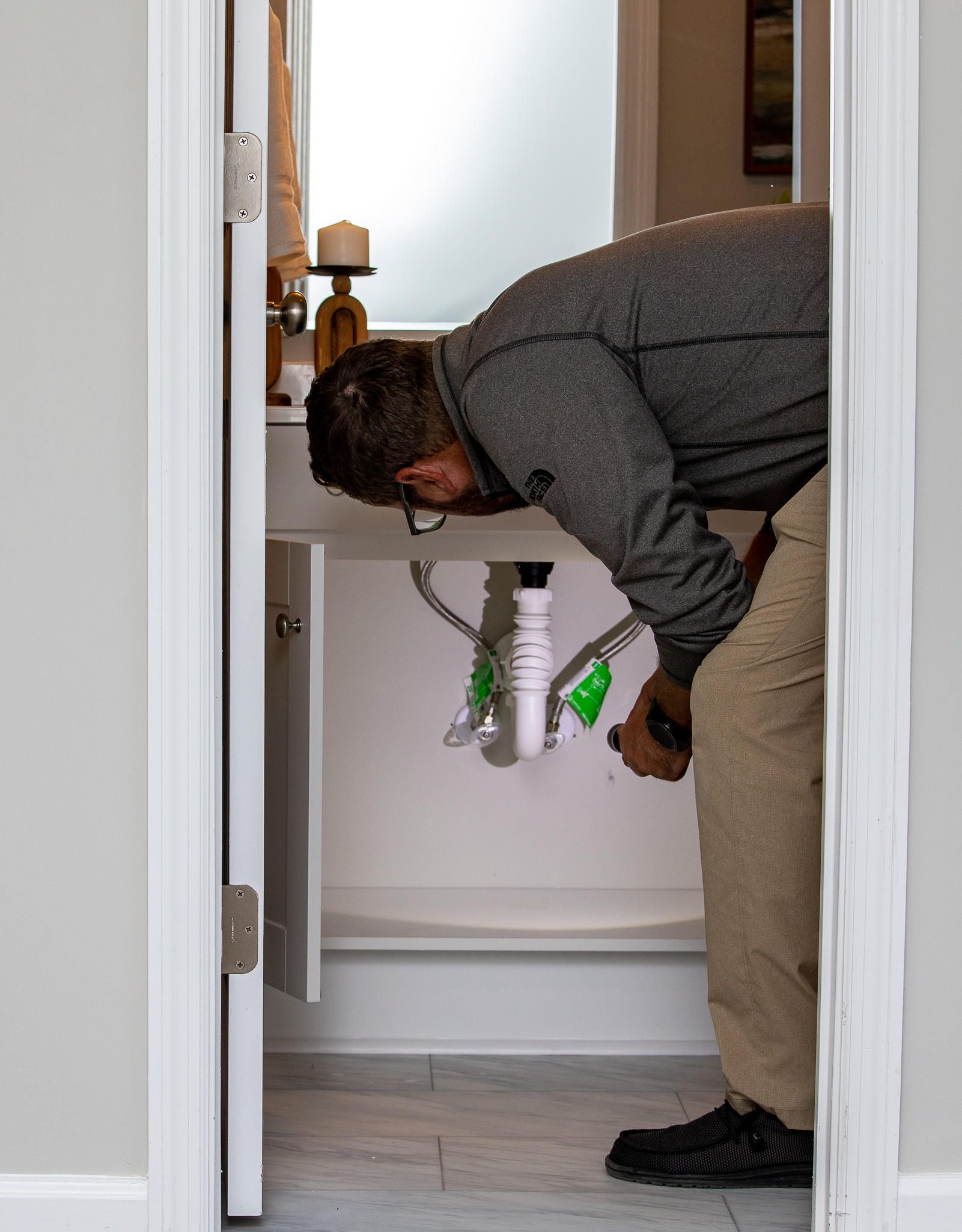 Man working on plumbing under a bathroom sink, seen through a doorway. White cabinet, grey wall, black shoes.