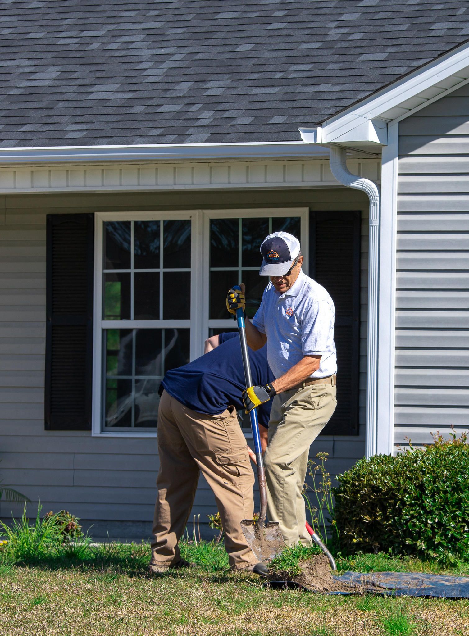 Two people digging in a yard near a house with a window, one using a shovel.