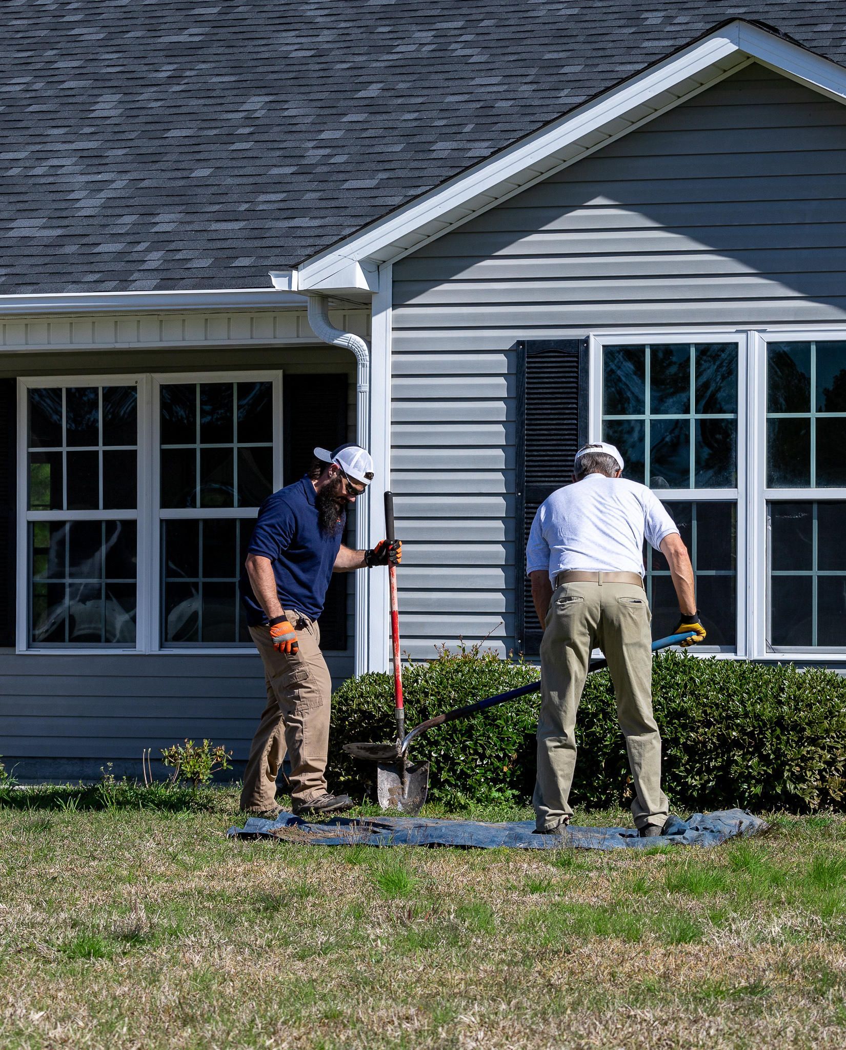 Two people digging in front of a house with a shovel. They are working on a lawn in front of windows.