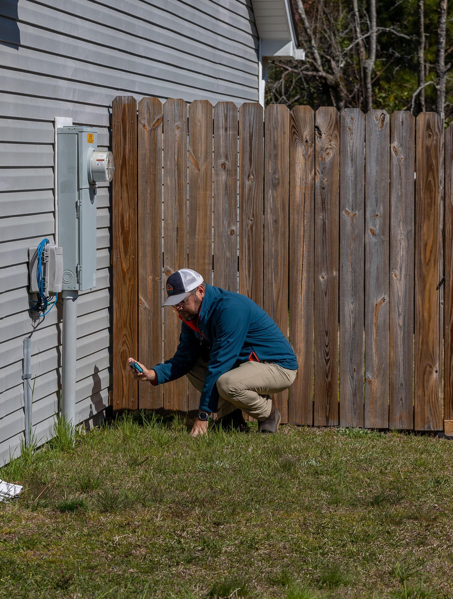 Person squats, checking a device near a utility box and wooden fence.