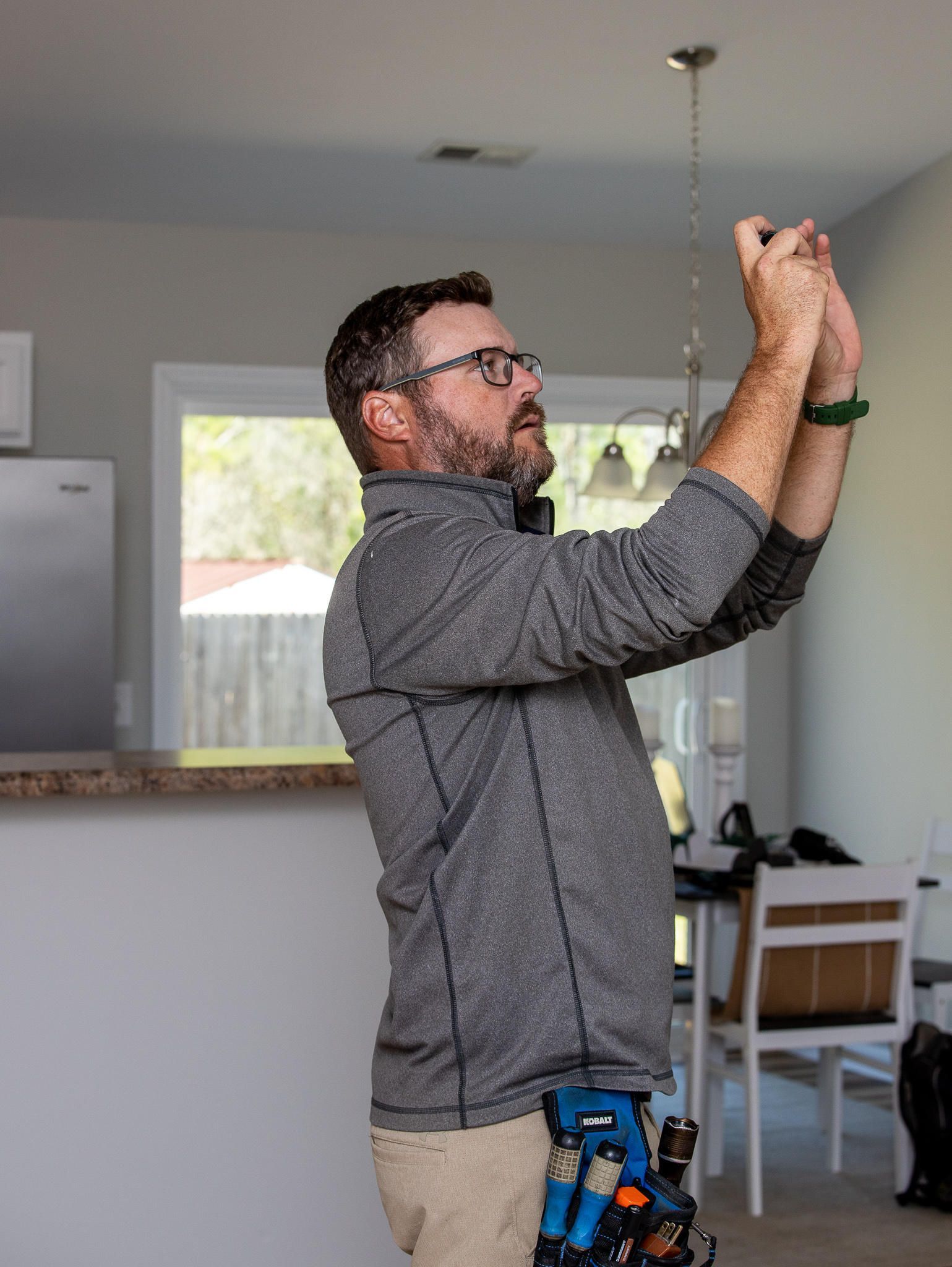 Man in glasses installing a light fixture in a house. He wears a gray jacket and tool belt.