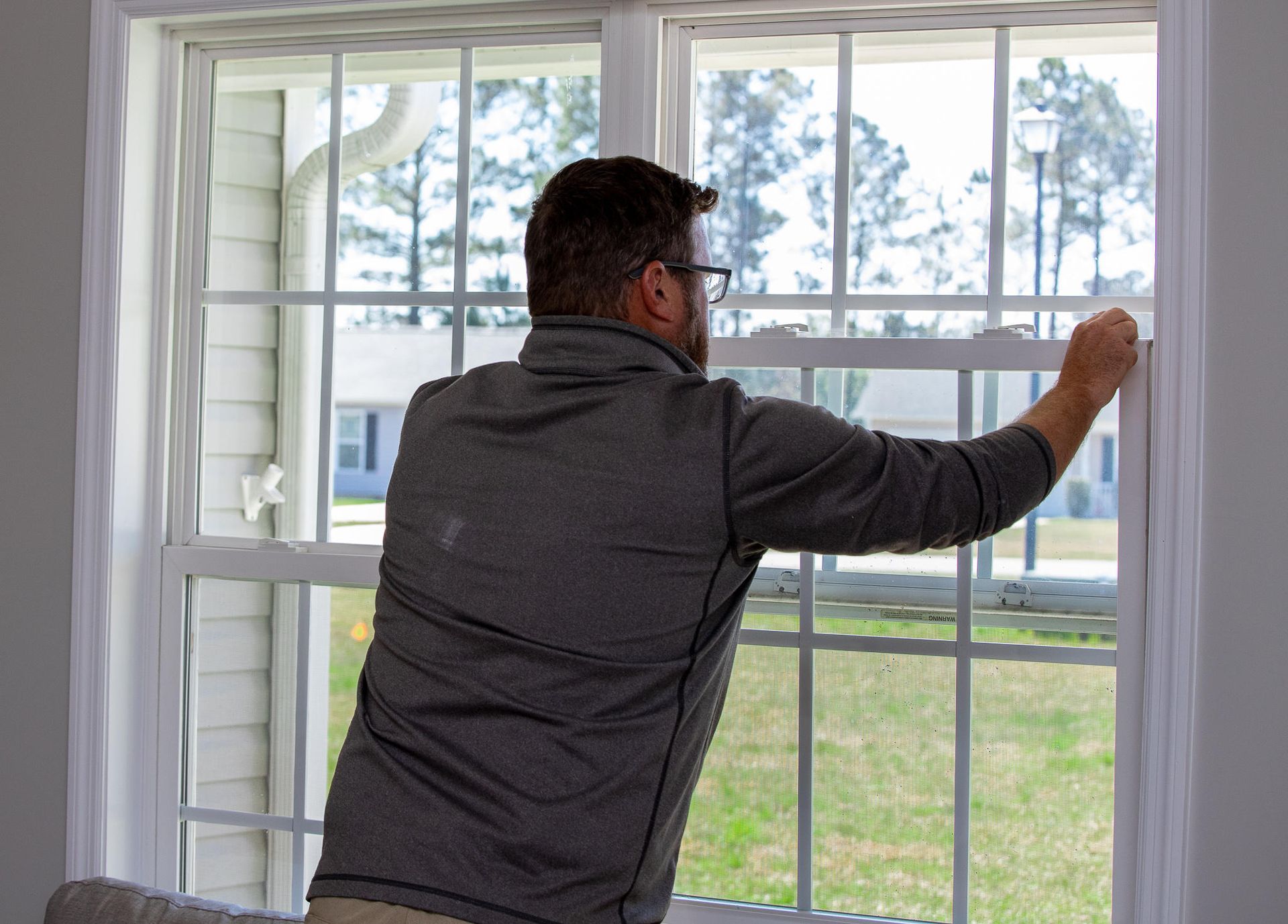 Man opening a white window with grid panes, looking outside. He's inside a room with a view of a lawn and house.