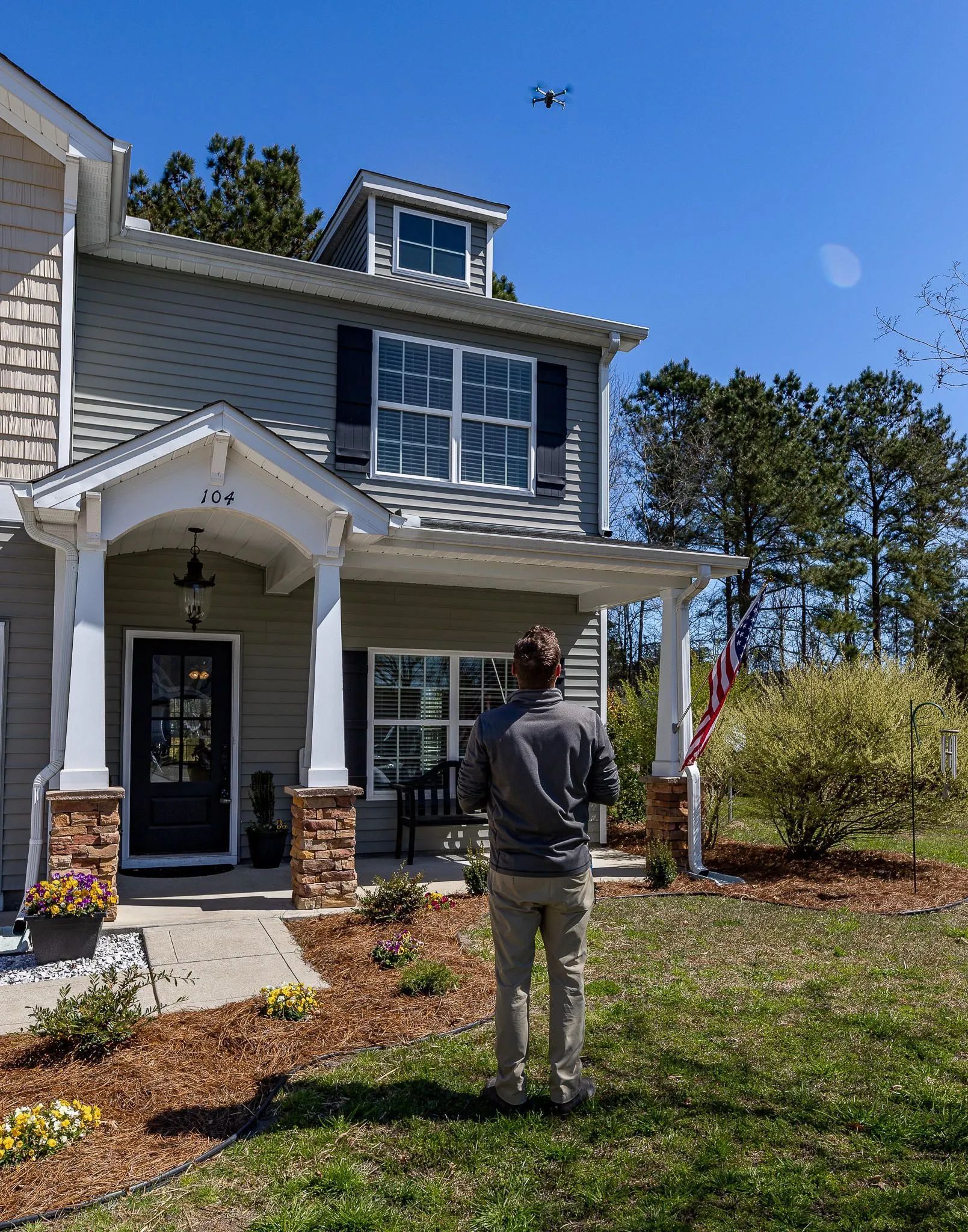 A person stands in a grassy yard, facing a two-story gray house, while operating a drone hovering in the clear blue sky.