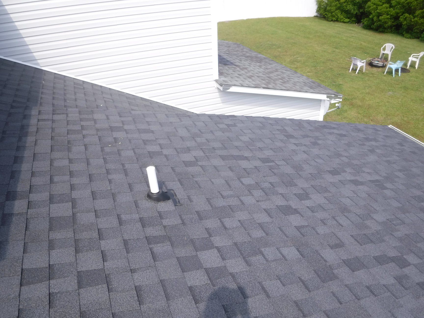 Dark gray shingle roof with a white pipe and white building next to green grass and patio furniture.