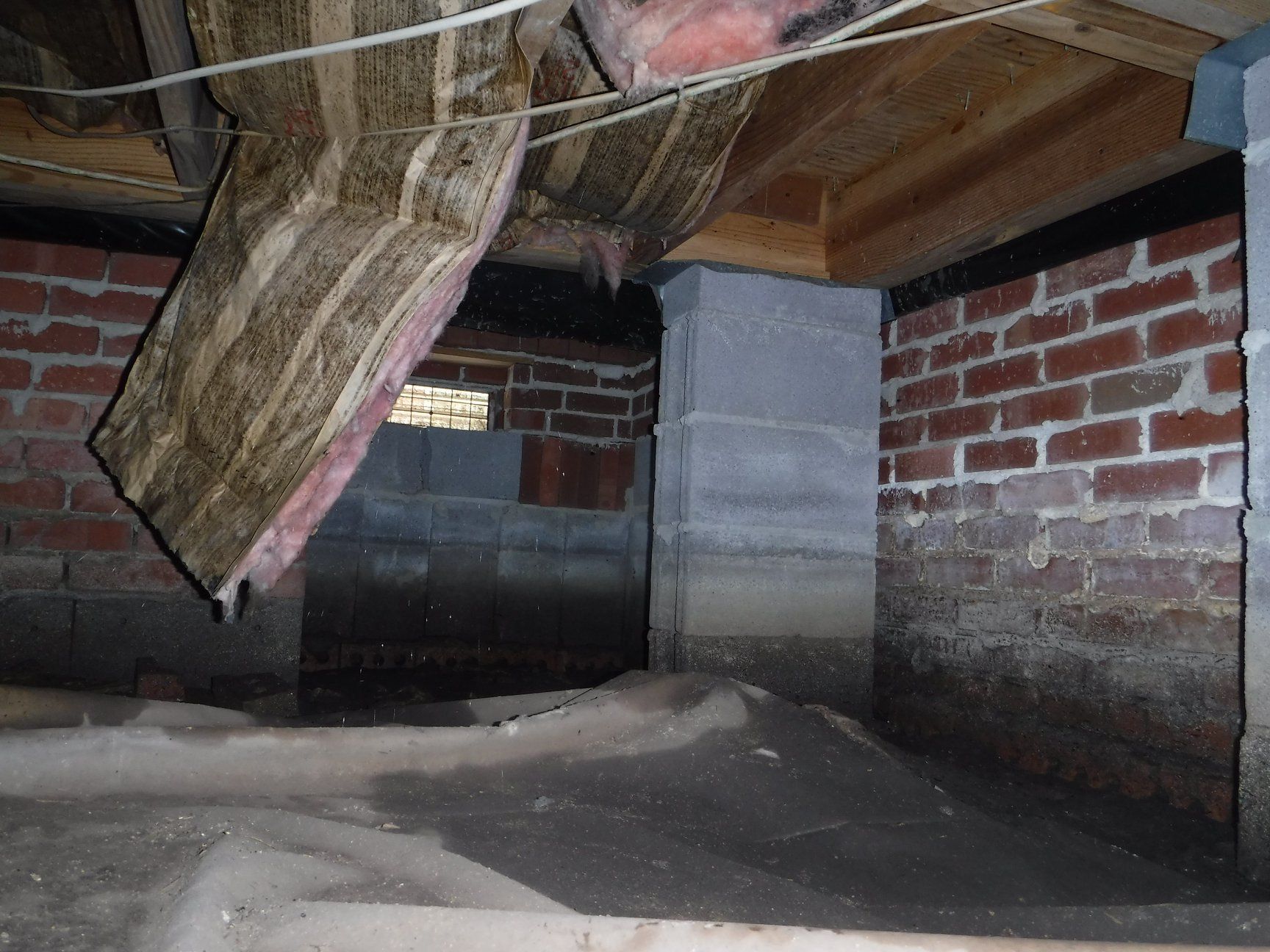Crawl space with brick walls, concrete pillar, and exposed wood framing. Dusty ground and debris present.