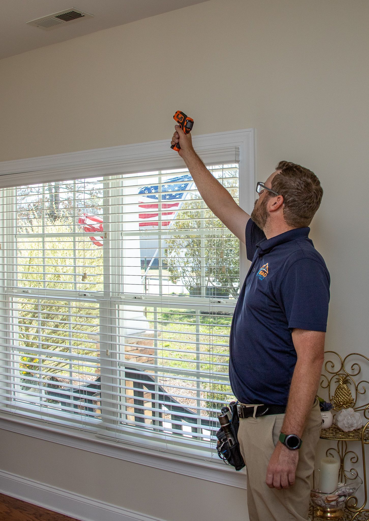 Man inspecting wall above a window. He's holding a tool, standing indoors. Blinds and a decorative shelf are visible.