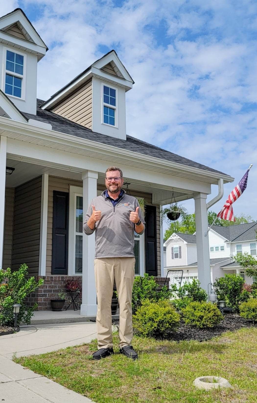 Man standing in front of a house, giving thumbs up. American flag visible. Sunny day.